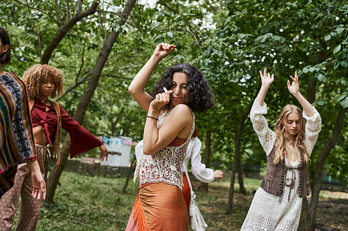 joyful multiracial woman in boho clothes looking at camera and dancing in retreat center