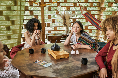 multiracial woman drinking tea near girlfriends and tarot cards on wooden table, women retreat
