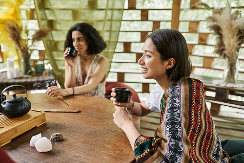 cheerful young woman with tea cup near multiracial girlfriend at wooden table in retreat center