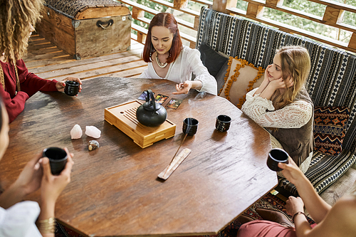 woman reading tarot cards near multiethnic girlfriends and tea on wooden table in cozy house