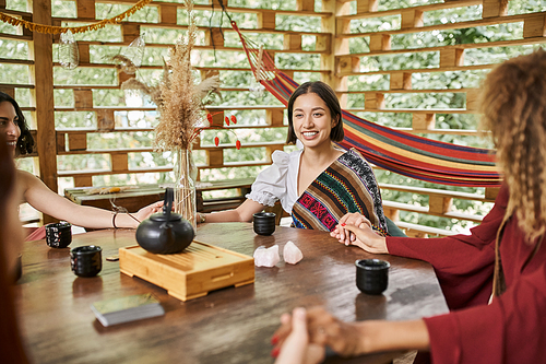 happy woman in boho style clothes meditating with girlfriends at table with tea in retreat center