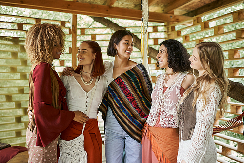 women retreat, happy multiethnic girlfriends in boho style outfit looking at each other in cottage