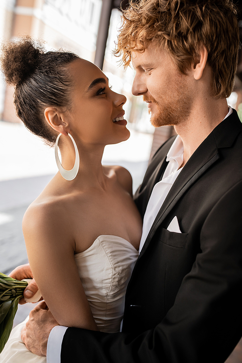 joyful and elegant interracial newlyweds looking at each other on street, wedding in urban setting