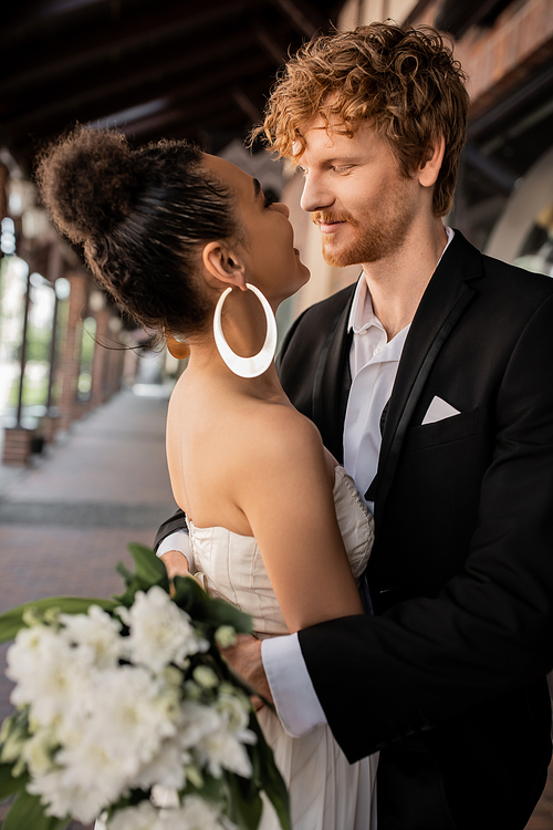 joyful interracial couple embracing and looking at each other, wedding attire, urban setting