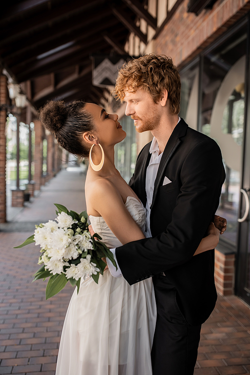 side view of stylish multiethnic man with flowers looking at african american bride on city street