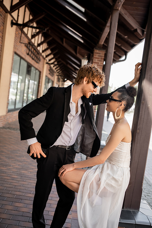 stylish redhead groom smiling near young african american bride on street, wedding in city