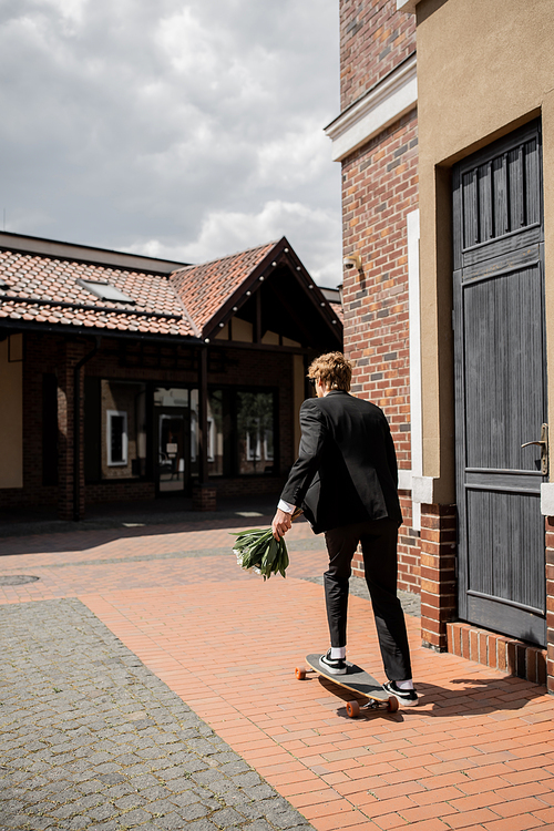 back view of groom in black suit holding wedding bouquet and  riding longboard on city street