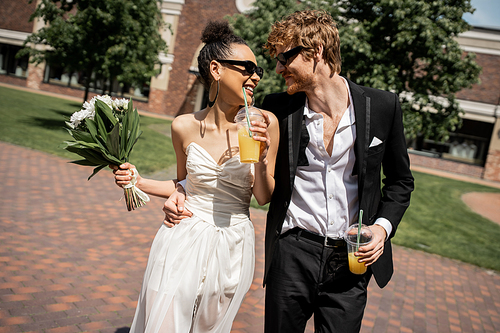 joyful multiethnic newlyweds in sunglasses walking with orange juice, wedding on urban street