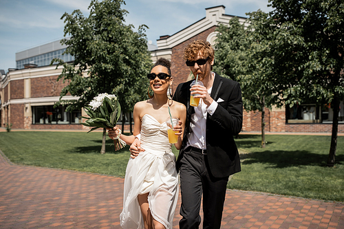 elegant newlyweds in sunglasses walking with orange juice and flowers in european city