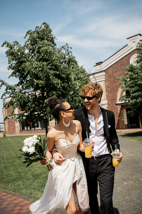 diverse couple with orange juice and flowers smiling at each other, wedding on street