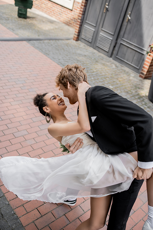 stylish redhead man embracing joyful african american bride, wedding on urban street