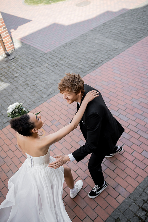 happy interracial couple in wedding attire running towards each other on urban street, top view