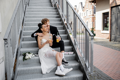 multiethnic newlyweds with orange juice sitting on stairs and looking away, wedding in city