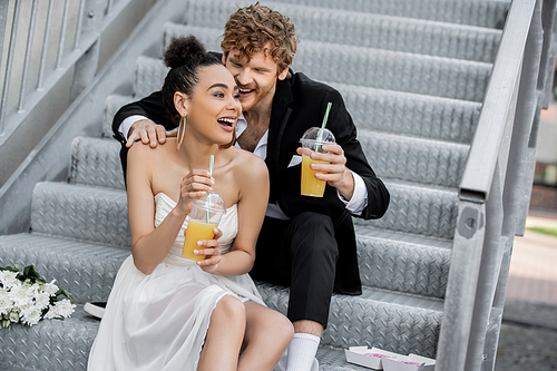 excited african american bride with orange juice laughing near groom on stairs outdoors