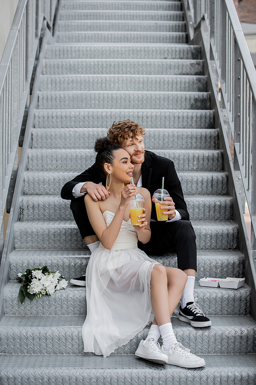 happy interracial newlyweds with orange juice looking away on street stairs near bouquet