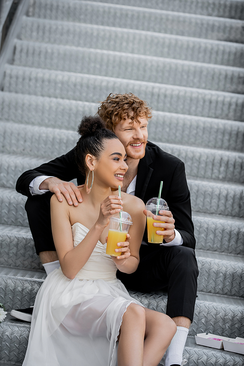 redhead groom embracing happy african american bride sitting with orange juice on stairs in city