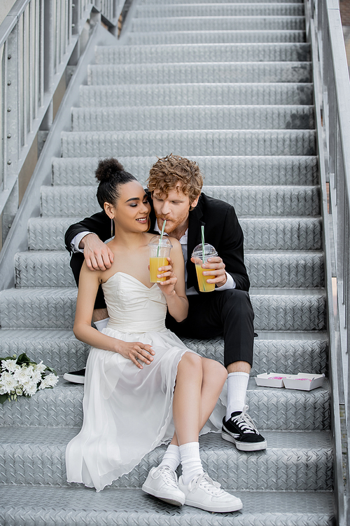 redhead man drinking orange juice from straw near african american bride, sitting on stairs in city