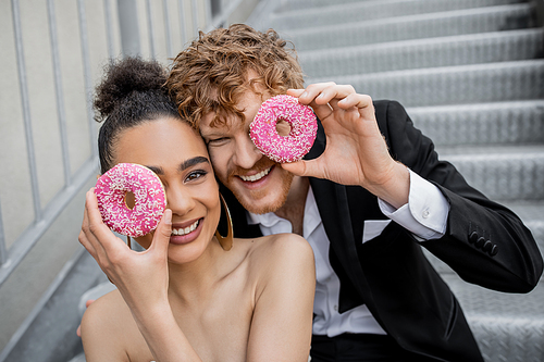 fun, happiness, wedding outdoors, excited multiethnic couple obscuring face with sweet donuts