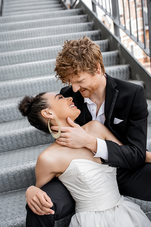 smiling redhead man hugging young african american bride on stairs in city, tenderness, happiness