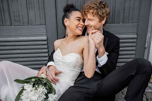 cheerful multiethnic newlyweds sitting near city building and holding hands, wedding attire, flowers