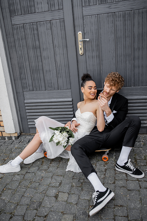 redhead man holding hands of african american bride with flowers, sitting on pavement near doors