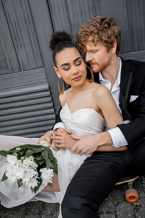 redhead groom hugging african american bride with closed eyes and flowers, sitting near building