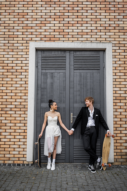 joyful interracial newlyweds with longboard and skateboard looking at each other near city building