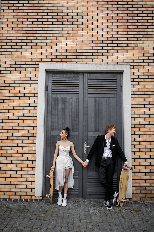 carefree multiethnic newlyweds with longboard and skateboard holding hands near city building