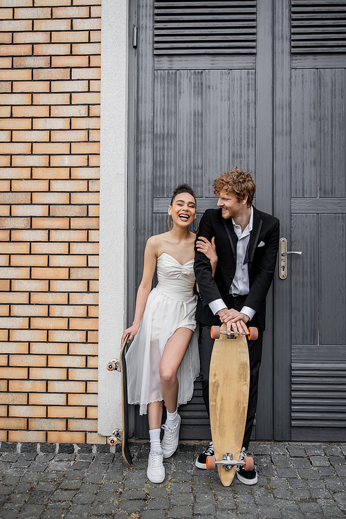 young interracial couple with skateboard and longboard near city building, happiness, laugh