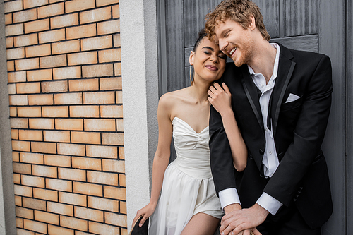 delighted african american bride embracing happy redhead groom near city building, closed eyes