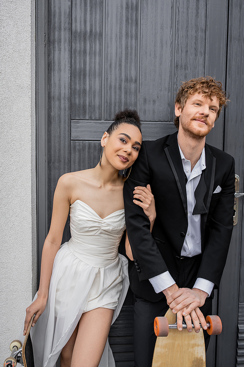 african american bride smiling at camera near redhead groom near building, longboard, skateboard