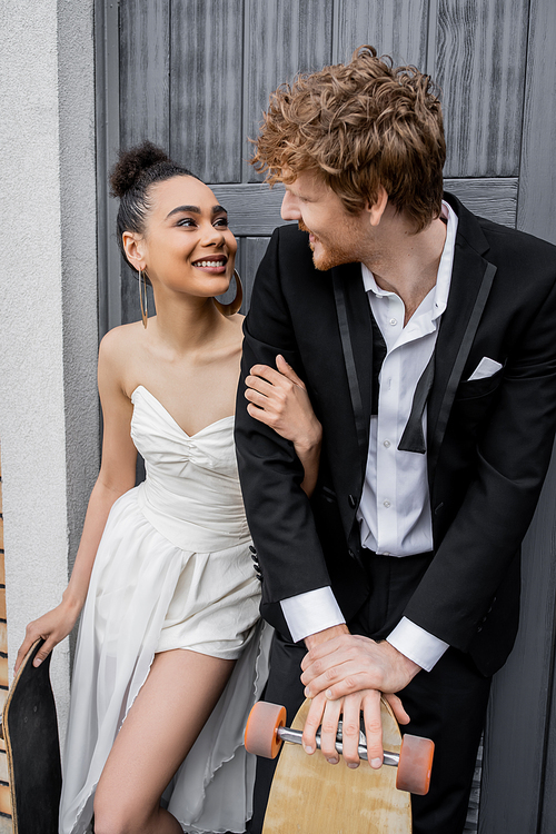 joyful multiethnic newlyweds with longboard and skateboard smiling at each other near city building