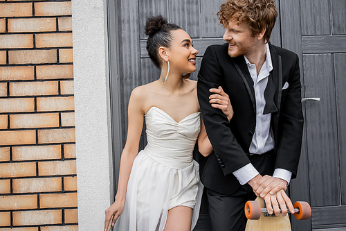 happy interracial newlyweds with longboard and skateboard smiling at each other near city building