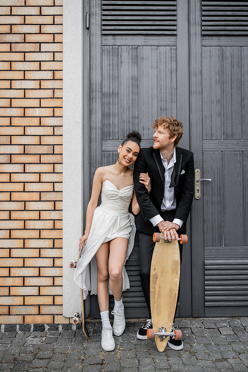 interracial couple in wedding attire, with longboard and skateboard near doors of city building