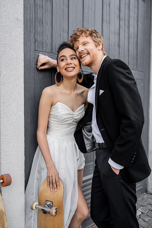 pleased redhead groom with hand in pocket, african american bride with skateboard near city building