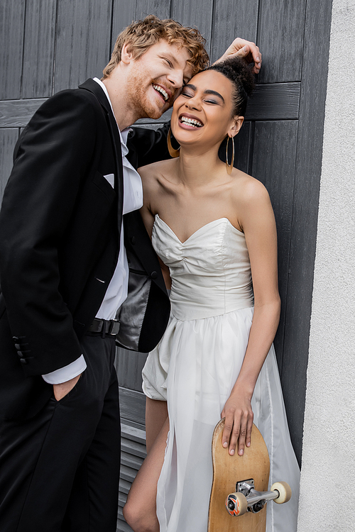 redhead groom and elegant african american bride with skateboard laughing near building in city