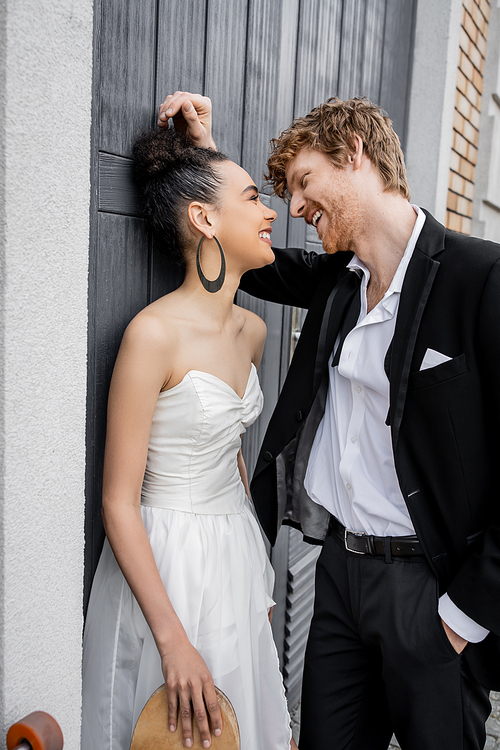 overjoyed groom with hand in pocket, stylish african american bride with skateboard near building