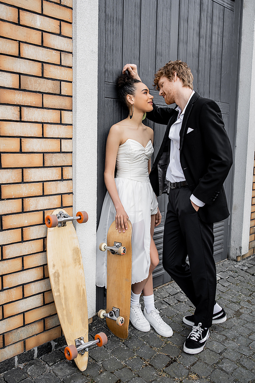 elegant redhead groom with hand in pocket near african american bride, longboard and skateboard