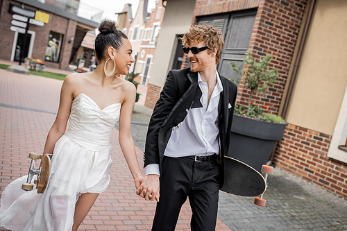 happy interracial couple with longboard and skateboard walking on street, wedding attire, sunglasses