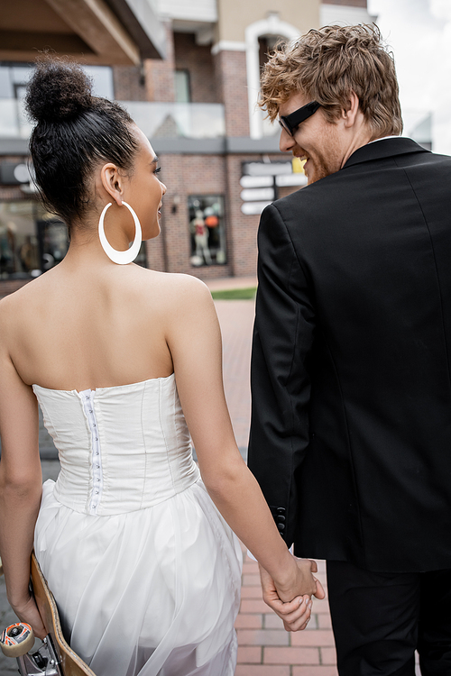 redhead groom in sunglasses, african american bride with longboard looking at each other on street