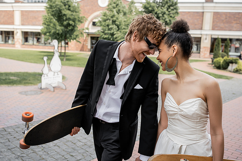 overjoyed multiethnic newlyweds with longboard and skateboard on street, sunglasses, wedding outfit