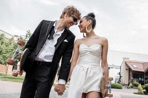 young interracial newlyweds with longboard and skateboard holding hands on street, happiness