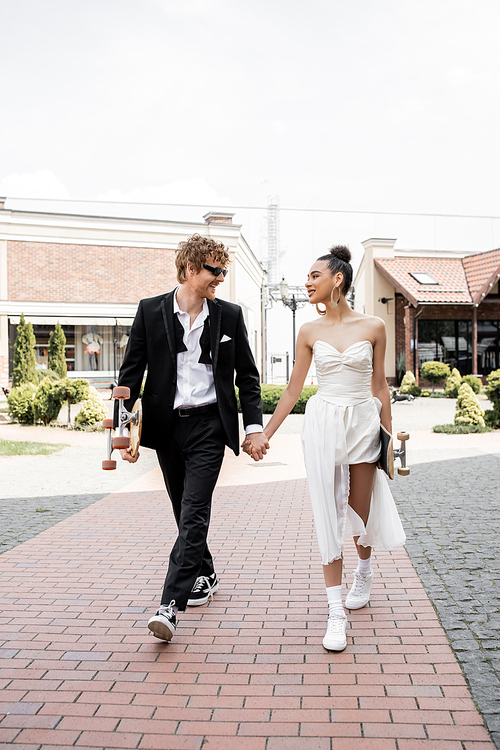 stylish interracial couple with longboard and skateboard holding, and walking in city, wedding day