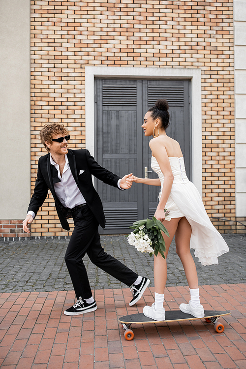 redhead groom in sunglasses holding hands with african american bride with bouquet riding longboard