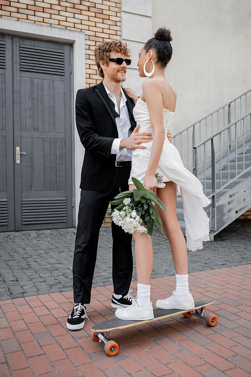 redhead groom in sunglasses hugging young african american bride with flowers standing on longboard