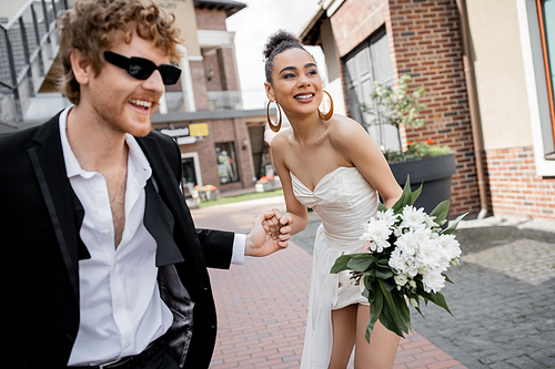 african american american bride with flowers and redhead groom in sunglasses holding hands on street