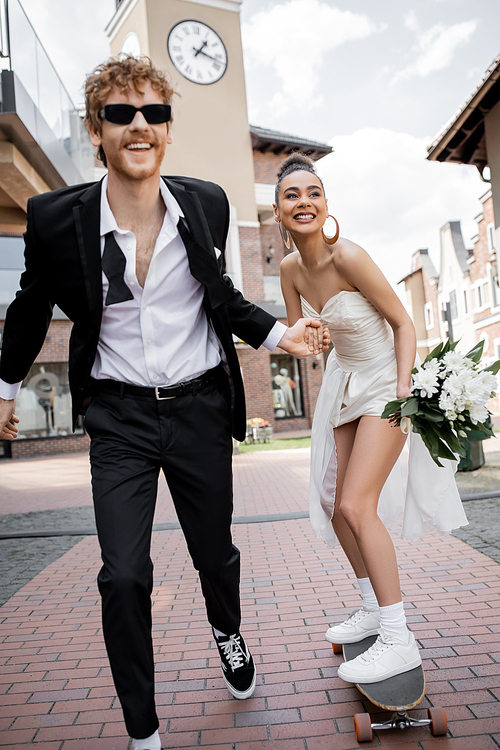 elegant african american woman in wedding dress riding longboard near groom in sunglasses