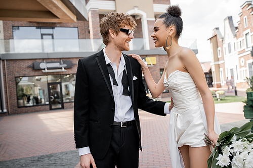 outdoor wedding, modern and stylish interracial newlyweds looking at each other on street