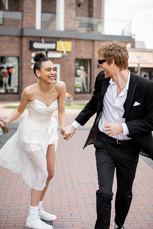 urban romance, elegant interracial newlyweds holding hands and running in european city