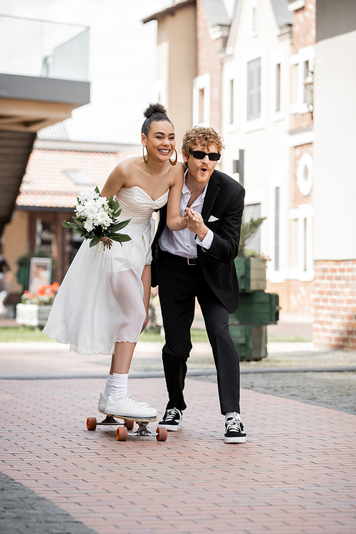 overjoyed african american woman in wedding dress riding longboard near elegant groom on street